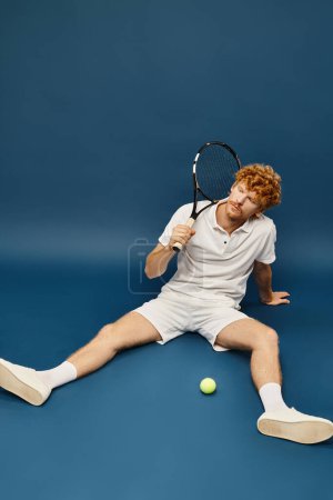 sportlicher Rotschopf in weißen trendigen Klamotten mit Schläger sitzt neben Tennisball auf blauem Hintergrund