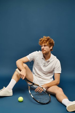 trendy redhead man in white tennis clothes with racquet sitting near tennis ball on blue backdrop