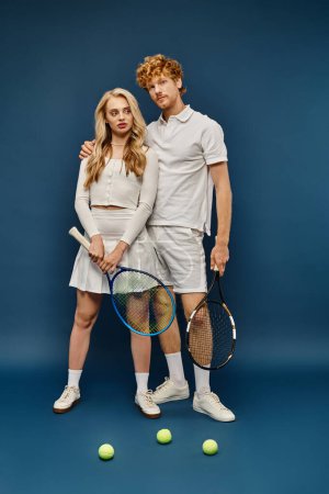 Photo for Full length of trendy couple in white tennis outfit posing with tennis racquets near balls on blue - Royalty Free Image