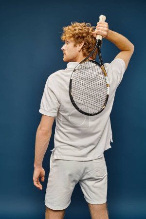 Rückansicht eines jungen rothaarigen Mannes im weißen Tennisoutfit, der mit Schläger auf blauer, zeitloser Mode posiert