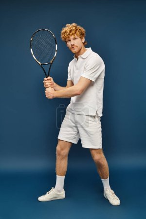 trendy schöner rothaariger Mann in weißer Kleidung spielt Tennis und schaut in die Kamera vor blauem Hintergrund