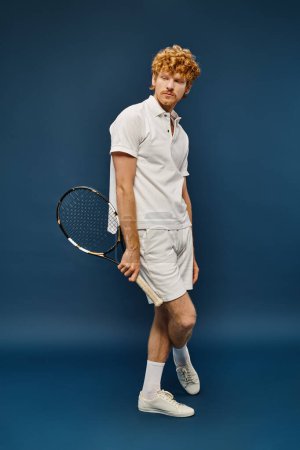 volle Länge des modischen rothaarigen Mannes im weißen Outfit mit Tennisschläger vor blauem Hintergrund