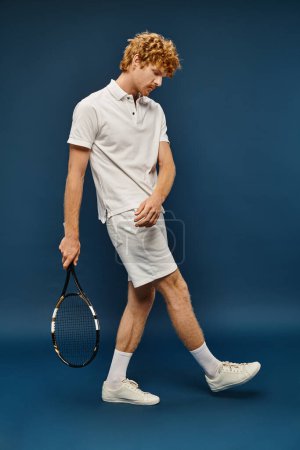 full length of young redhead man in white trendy outfit walking with tennis racquet on blue backdrop