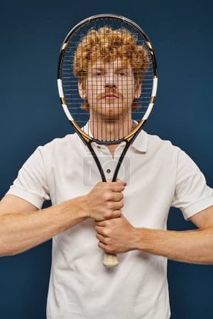 Photo for Trendy redhead man in white clothes obscuring face with tennis racquet and looking at camera on blue - Royalty Free Image