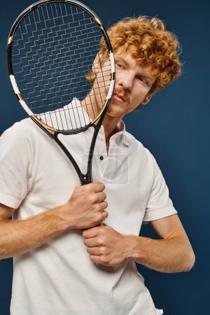 schöner rothaariger Mann im weißen Tennisoutfit mit Schläger vor dem Gesicht auf blauer, zeitloser Mode