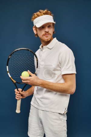 Sportlicher Mann in weißem Tennisoutfit und Sonnenblende mit Schläger und Ball auf blauem, wohlhabendem Sport