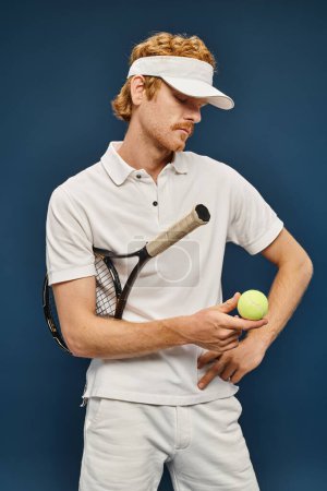 junger rothaariger Mann in weißer Kleidung und Sonnenblende mit Schläger und Blick auf Tennisball auf blau