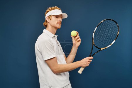 junger hübscher rothaariger Mann in weißer Kleidung und Sonnenblende mit Schläger und Tennisball auf blauem Grund