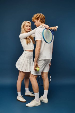 Photo for Stylish blonde woman with tennis racquet embracing redhead man in white attire on blue, full length - Royalty Free Image