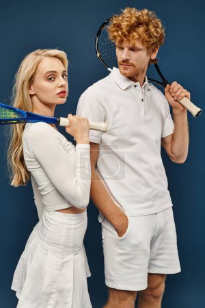 Photo for Handsome redhead man looking at blonde woman with tennis racquet looking at camera on blue - Royalty Free Image