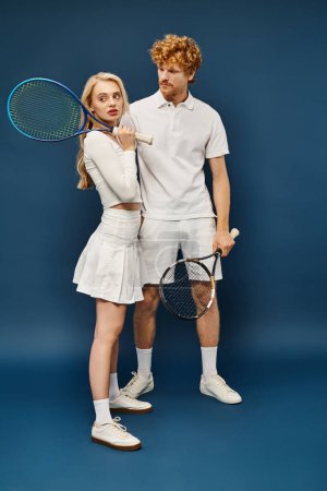 Photo for Full length of redhead man looking at blonde woman with tennis racquet looking away on blue - Royalty Free Image