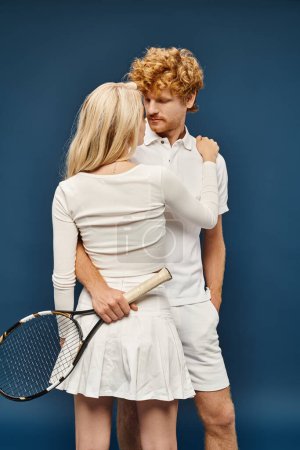 trendy blonde woman embracing young redhead man in white clothes with tennis racquet on blue