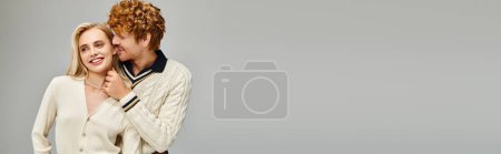 cheerful redhead man touching neck of smiling blonde woman in stylish clothes on grey, banner