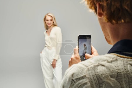 young man taking photo of fashionable blonde woman on grey backdrop, classic fashion concept