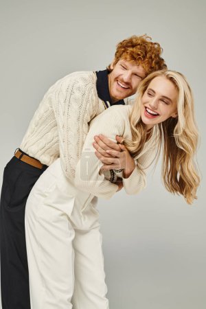 freudiger rothaariger Mann umarmt lachende blonde Frau und hat Spaß auf grauem Hintergrund, klassische Mode