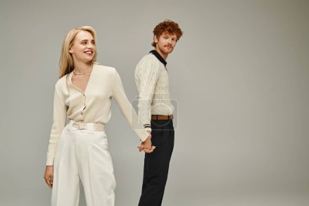 happy blonde woman and redhead man in trendy casual clothes holding hands on grey, old fashion style