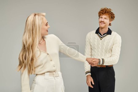 cheerful blonde woman and stylish redhead man holding hand on grey, old money style concept