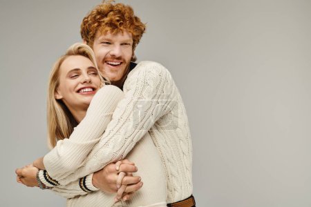 excited redhead man embracing blonde woman in trendy casual clothes on grey, timeless fashion