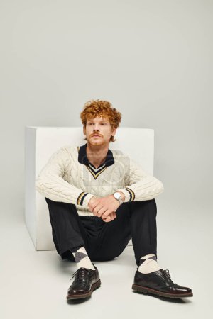 Foto de Hombre de ensueño con estilo en traje casual mirando hacia otro lado sentado cerca del cubo blanco en gris, concepto de moda - Imagen libre de derechos