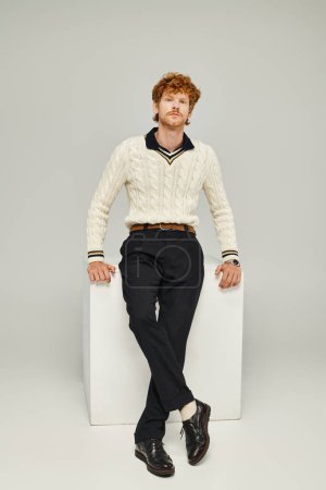 Foto de Hombre pelirrojo de moda en ropa casual sentado en cubo blanco y mirando a la cámara en gris - Imagen libre de derechos