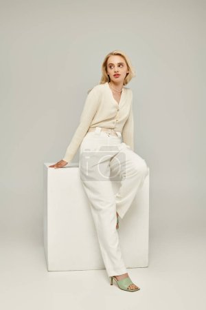 mujer rubia de moda en ropa ligera sentada en cubo blanco y mirando hacia otro lado en el fondo gris