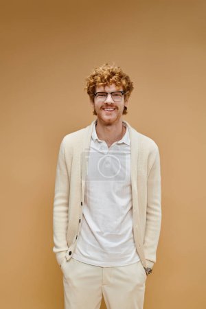 Foto de Hombre pelirrojo feliz en gafas y ropa ligera con las manos en los bolsillos mirando a la cámara en beige - Imagen libre de derechos