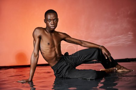 appealing african american man sitting topless on water surface and looking at camera, lights