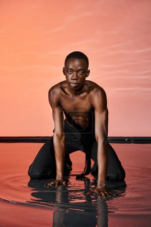 alluring african american man in wet pants sitting on water surface and looking at camera, red light