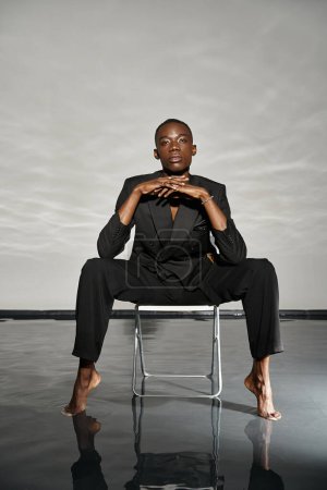 Téléchargez les photos : Séduisant afro-américain en costume déboutonné sexy assis sur la chaise et regardant la caméra - en image libre de droit