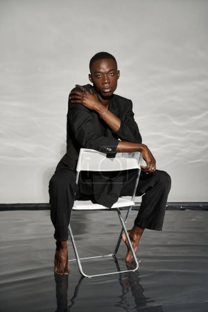 Téléchargez les photos : Attrayant sexy afro-américain en élégant costume noir assis sur la chaise et regardant la caméra - en image libre de droit