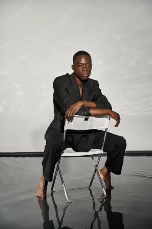 Téléchargez les photos : Bel homme sexy afro-américain en costume noir élégant assis sur la chaise et regardant la caméra - en image libre de droit