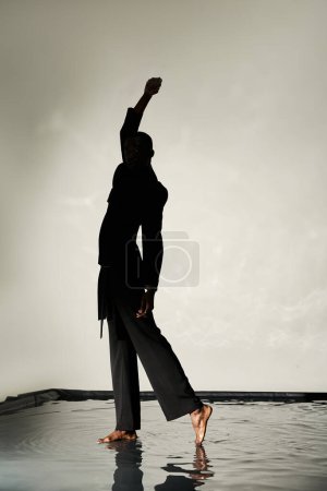 silhouette of trendy african american man posing barefoot in suit on gray watery backdrop in shadows