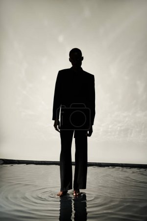 Silhouette eines verführerischen afrikanisch-amerikanischen Mannes, der barfuß im Anzug vor nasser Kulisse im Schatten posiert