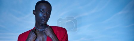 trendy african american man in red vibrant blazer posing on blue watery background, banner