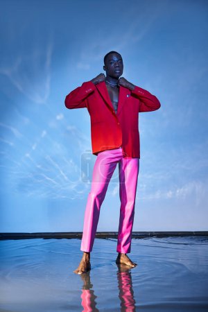 Téléchargez les photos : Engageant homme afro-américain élégant en pantalon rose et blazer rouge posant sur fond bleu aqueux - en image libre de droit