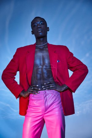 Téléchargez les photos : Engageant homme afro-américain élégant en pantalon rose et blazer rouge posant sur fond bleu aqueux - en image libre de droit