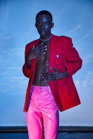 Téléchargez les photos : Charmant homme afro-américain élégant en pantalon rose et blazer rouge posant sur fond bleu aqueux - en image libre de droit