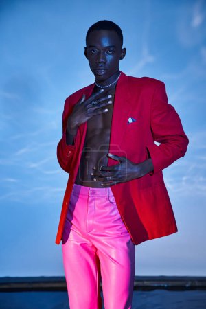 Téléchargez les photos : Bel homme élégant afro-américain en pantalon rose et blazer rouge posant sur fond bleu aqueux - en image libre de droit