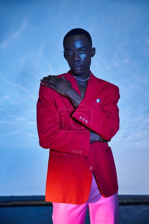 Téléchargez les photos : Attrayant homme afro-américain élégant en pantalon rose et blazer rouge posant sur fond bleu aqueux - en image libre de droit