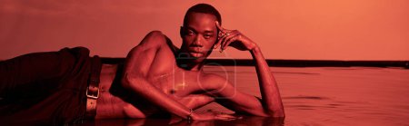 Foto de Atractivo hombre afroamericano posando en topless y reclinado en la superficie del agua en luces rojas, pancarta - Imagen libre de derechos