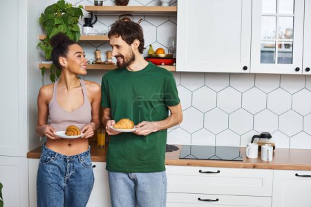 diversa pareja alegre en acogedora ropa de casa celebración de platos con deliciosos croissants mientras que en la cocina