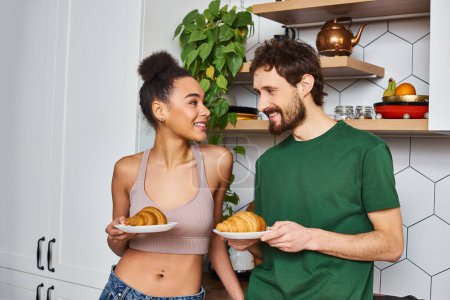 jolly interracial couple in casual outfits preparing to eat delicious croissants for breakfast