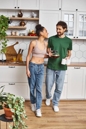 cheerful interracial couple in homewear preparing to drink hot coffee and smiling at each other