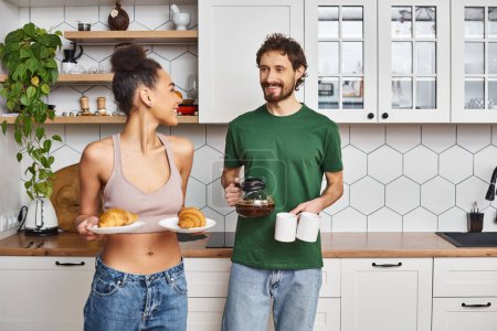 fröhliches multiethnisches Paar in Hauskleidung, das sich auf das Frühstück mit Kaffee und Croissants vorbereitet
