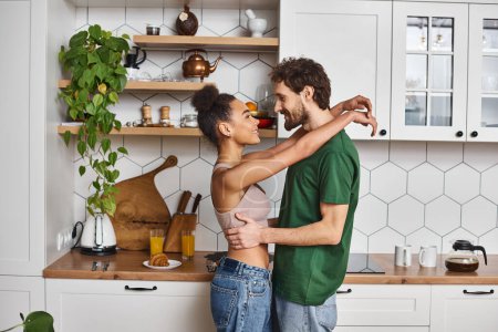 liebevolles fröhliches multikulturelles Paar in Hauskleidung, das sich herzlich umarmt und sich in der Küche amüsiert
