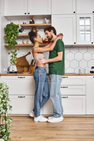 alegre pareja interracial atractiva en atuendos casuales abrazándose amorosamente antes de desayunar