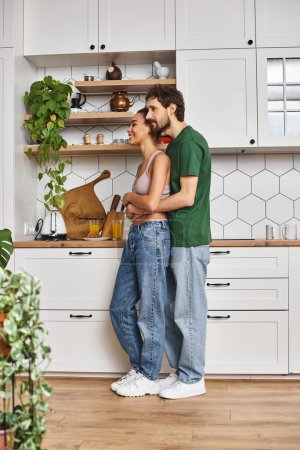ansprechendes fröhliches, vielfältiges Paar in lässiger Kleidung, das sich in der heimischen Küche herzlich umarmt