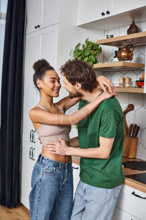 appealing jolly diverse couple in casual outfits hugging lovingly before eating breakfast at home