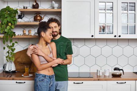 verführerisch gut aussehendes, multiethnisches Paar in lässiger Kleidung, das sich zu Hause in der Küche liebevoll umarmt
