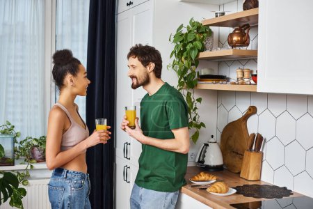 beautiful joyous diverse couple in homewear drinking fresh orange juice and smiling happily